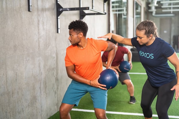 Two people performing a rotational medicine ball exercise with an Exos coach guiding form.