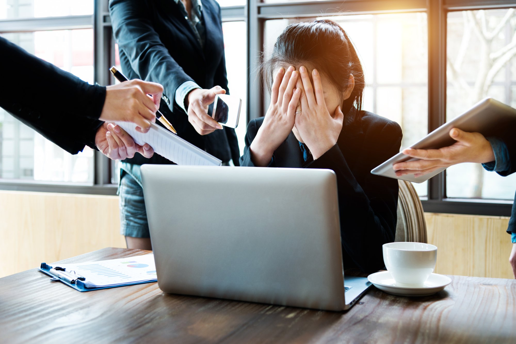 Stressed office worker covering face at desk while surrounded by multiple hands offering documents and a phone.