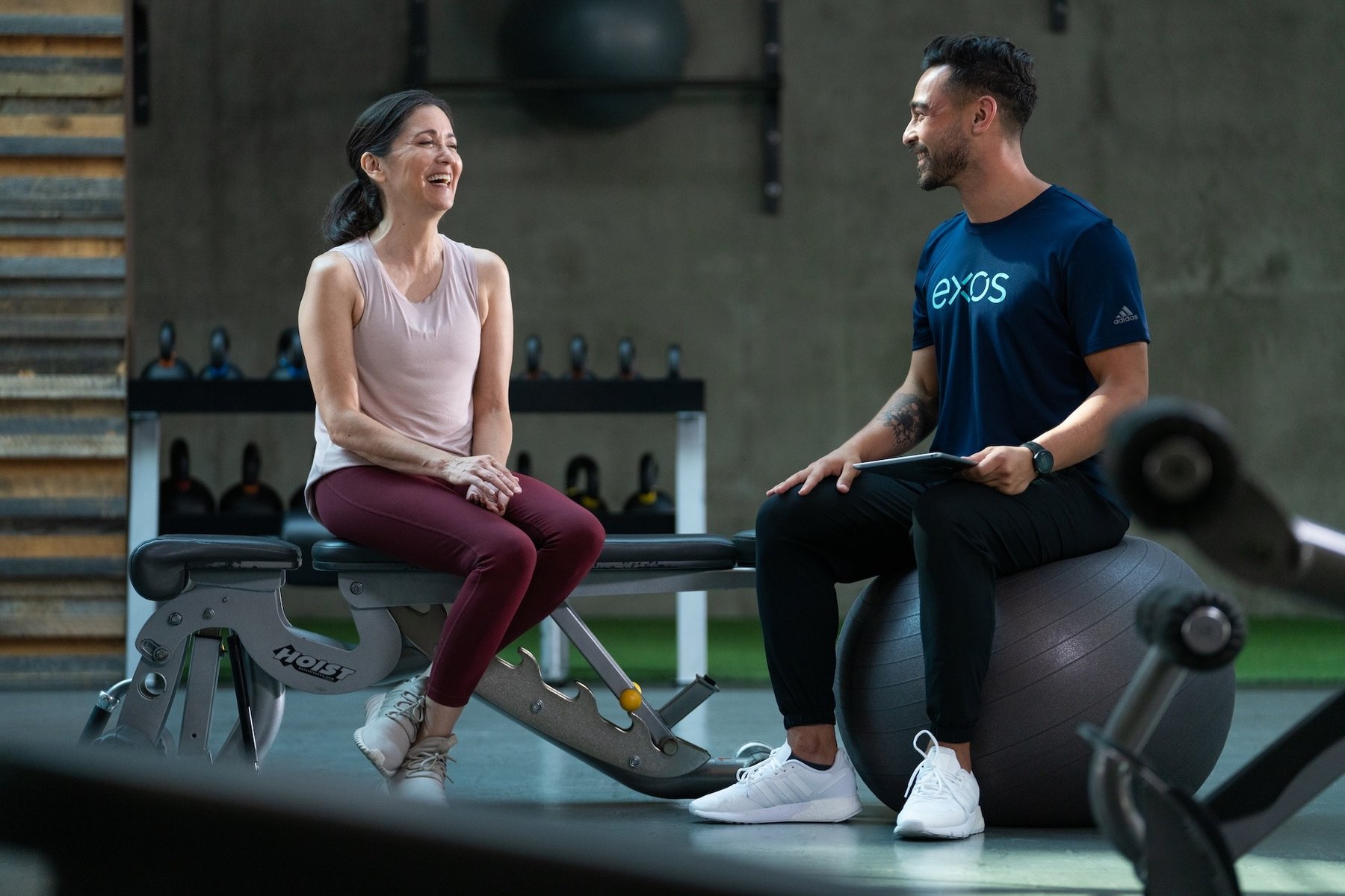 Woman in workout wear laughing with Exos trainer who holds tablet, both seated in modern fitness facility.