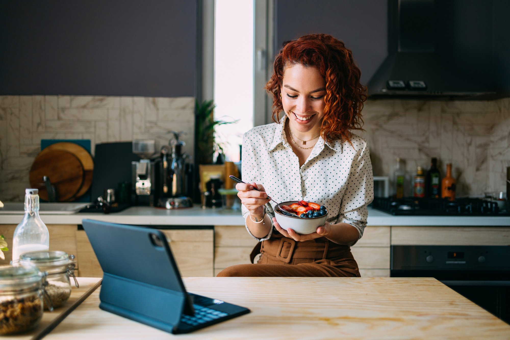 A woman with curly red hair smiles while eating from a bowl in a modern kitchen, with a laptop open on the counter.