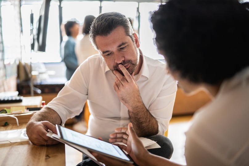Man in a white shirt attentively listening to a colleague discussing something on a tablet in a well-lit office setting.