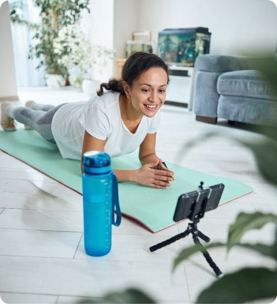 Smiling person holding a plank on a yoga mat at home, engaging in hybrid coaching through the Exos app on a smartphone.