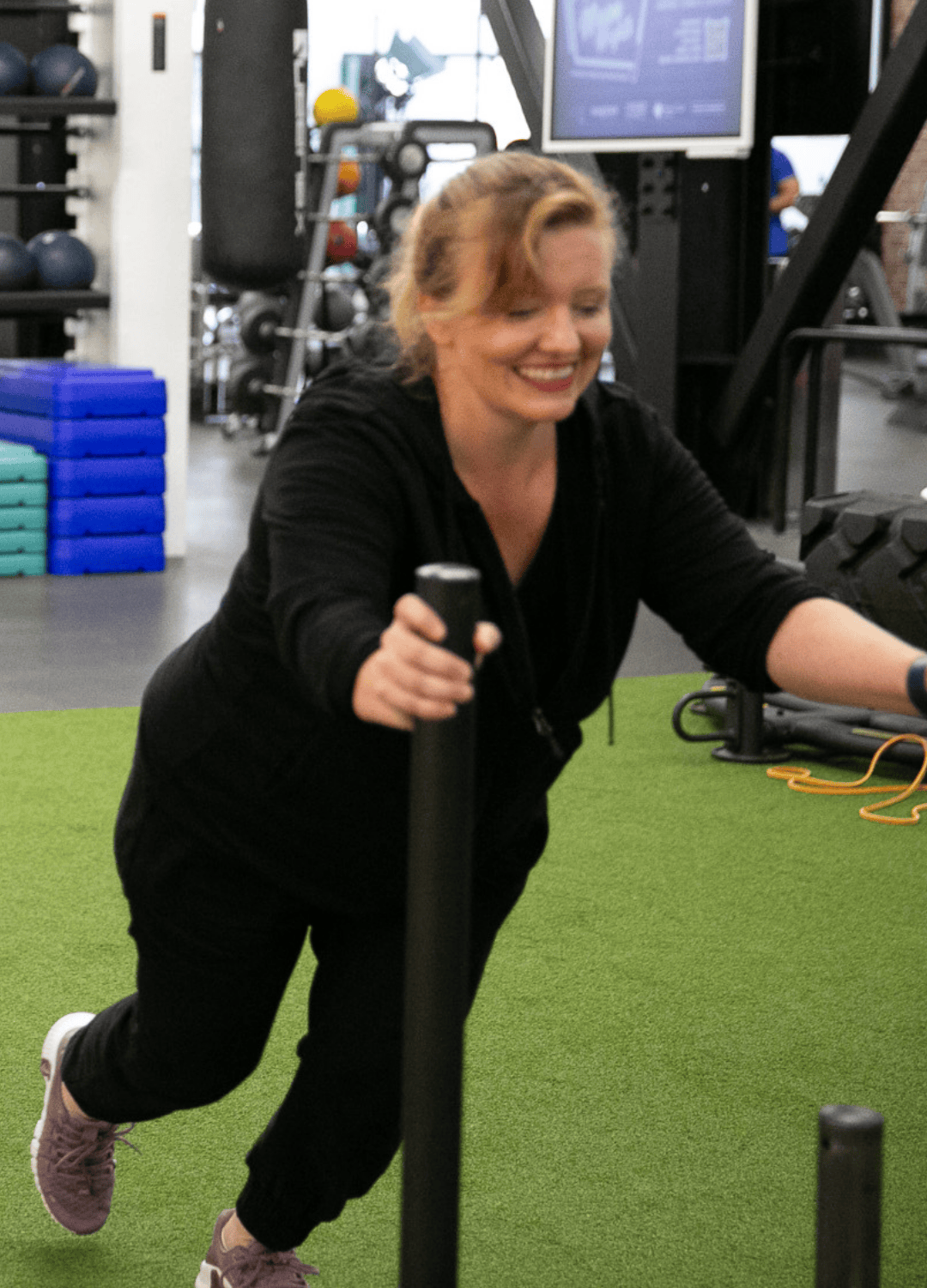 Woman smiles while pushing a weighted sled on turf in a gym, showing effort and enjoyment.