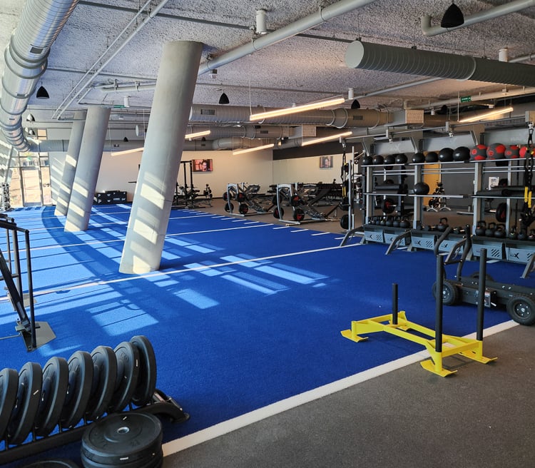 Functional training area with blue turf, sleds, bumper plates, and medicine balls in a bright, modern gym.