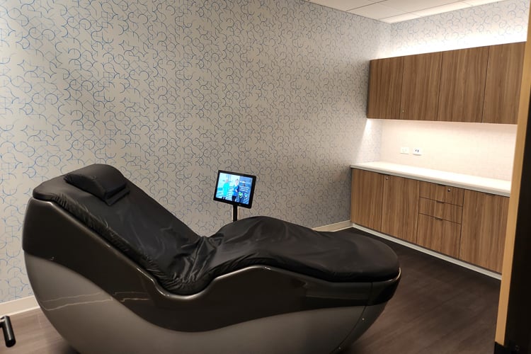 Private wellness room with a black massage chair, touchscreen display, and wood cabinetry.