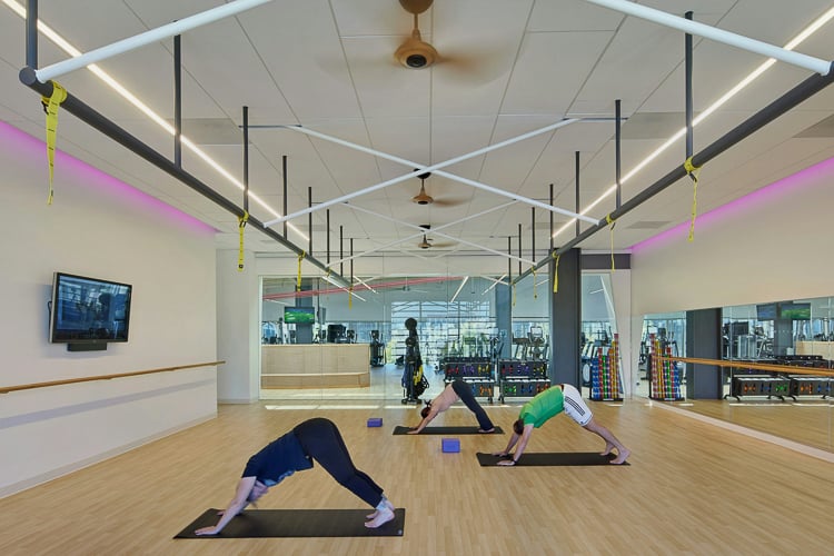 Three people in a fitness studio doing yoga poses on mats, with mirrored walls and bright lighting.