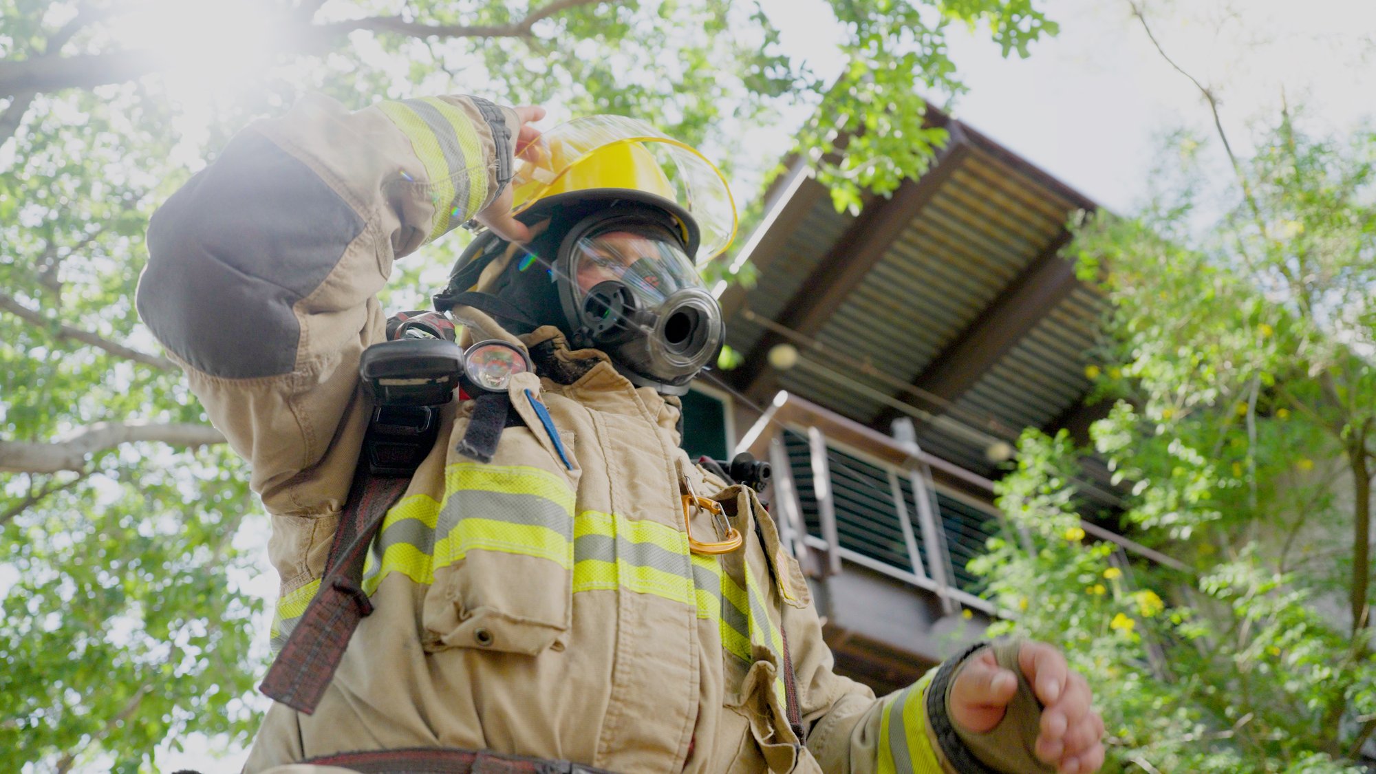 A firefighter adjusts his helmet under a tree in sunlight