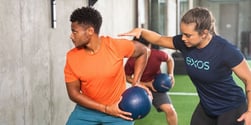 Two people performing a rotational medicine ball exercise with an Exos coach guiding form.