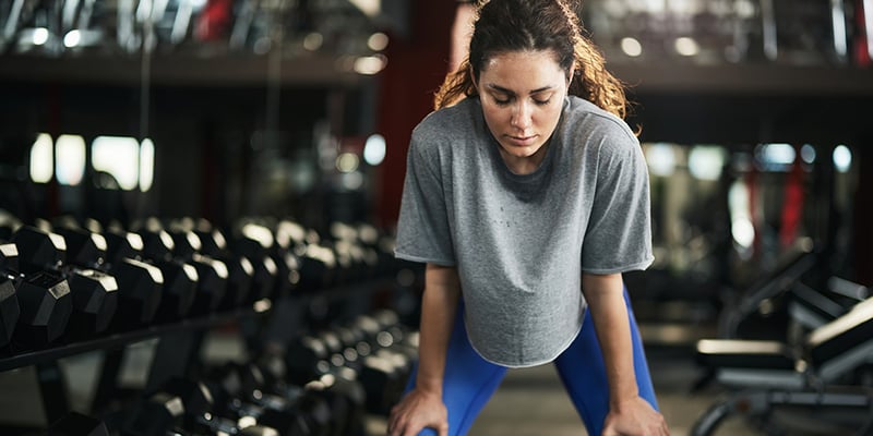 A woman in a gym leans forward, catching her breath after an intense workout, surrounded by rows of dumbbells.