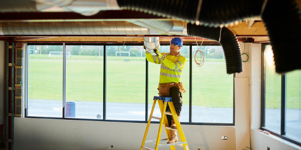 Construction worker on a ladder builds out air conditioning in an in-progress fitness center.
