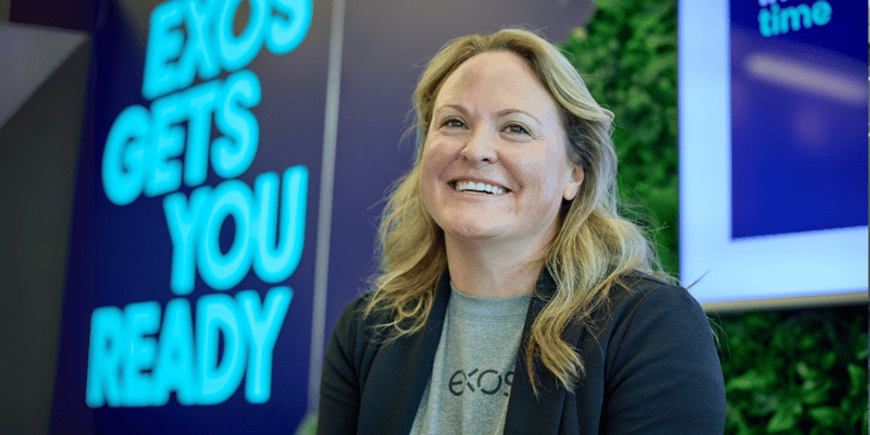 Smiling Exos team member stands before a vibrant “Exos Gets You Ready” sign, symbolizing our human-first performance coaching.