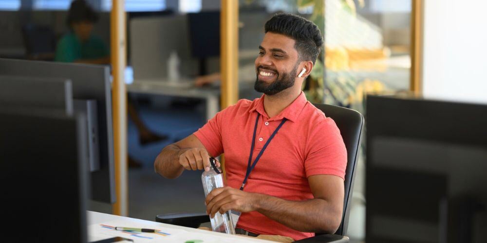 Smiling man in a red polo shirt opens a water bottle at his office desk, wearing wireless earbuds and a lanyard.