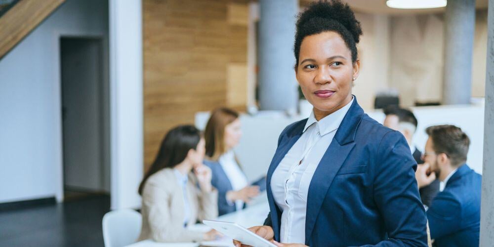 Confident businesswoman in a navy suit holding a tablet, standing in a modern office with colleagues collaborating in the background.