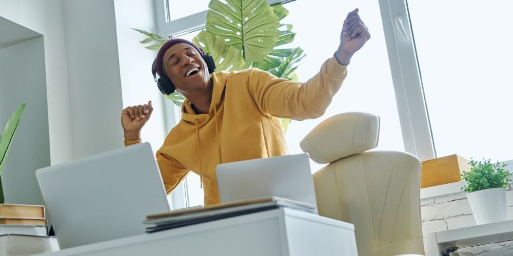 Man wearing headphones and a yellow hoodie stretches his arms at his desk.