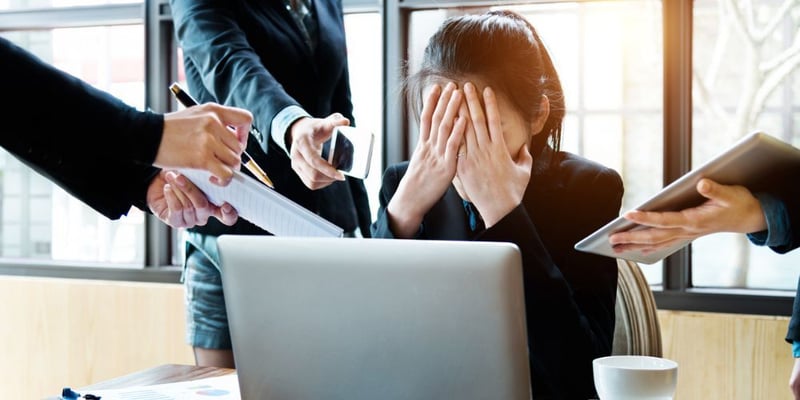 Stressed office worker covering face at desk while surrounded by multiple hands offering documents and a phone.