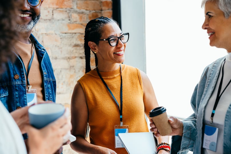 Three professionals smile and chat while holding coffee cups during a casual workplace gathering.