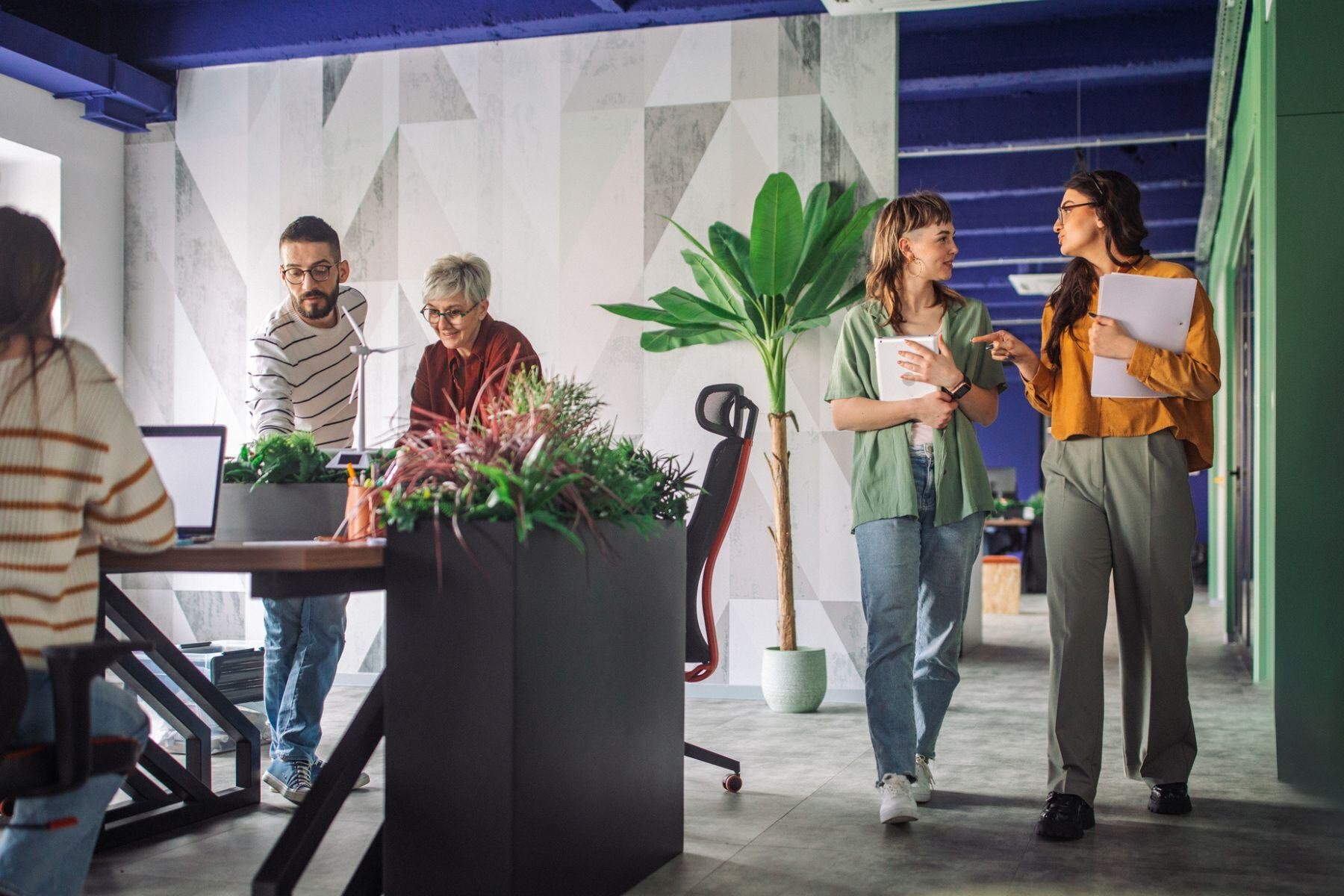 Modern office space with diverse professionals collaborating; two women walk and discuss while others work at desks with greenery.