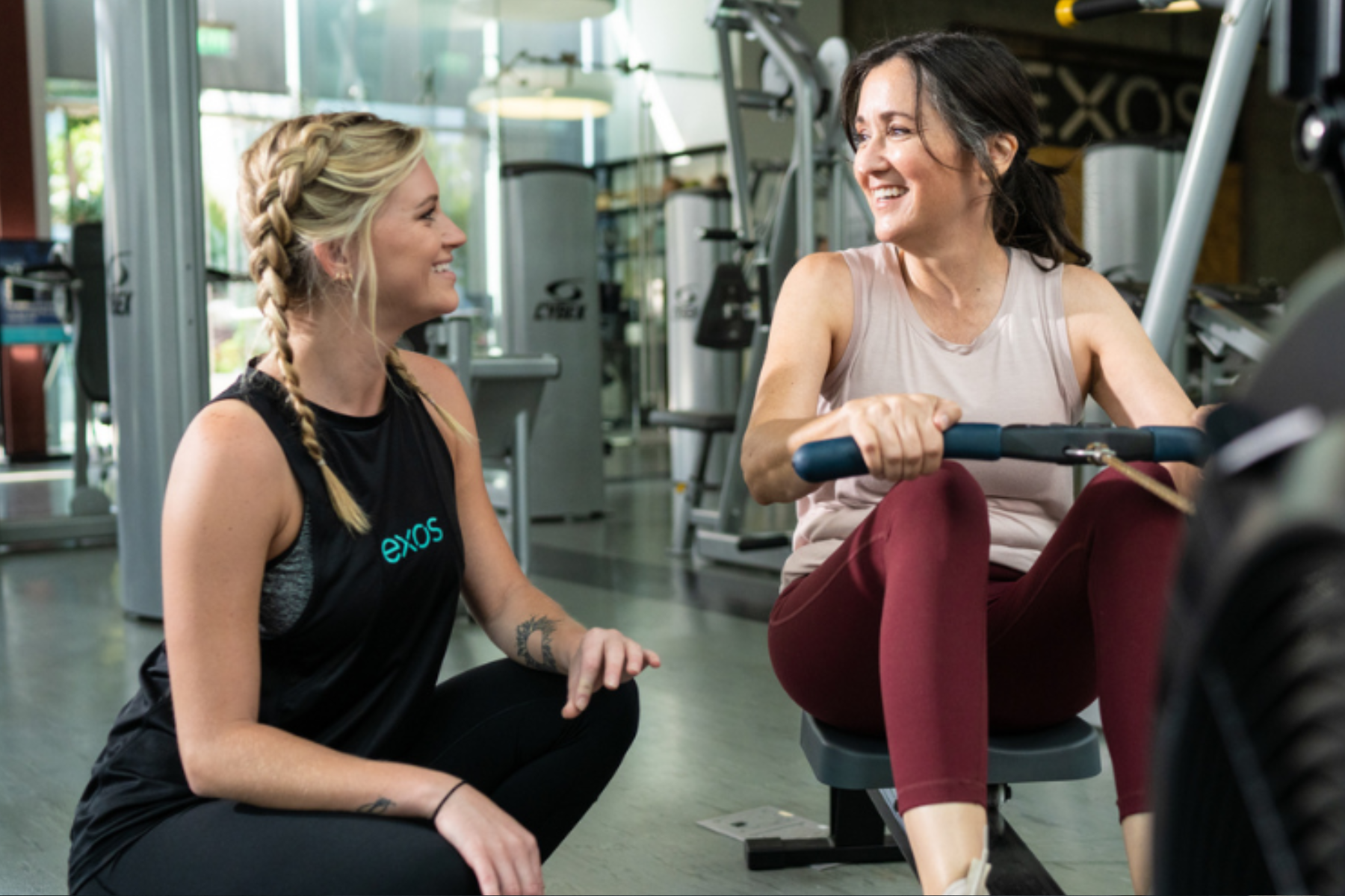 An Exos coach gives encouragement and advice to a woman on a rowing machine.