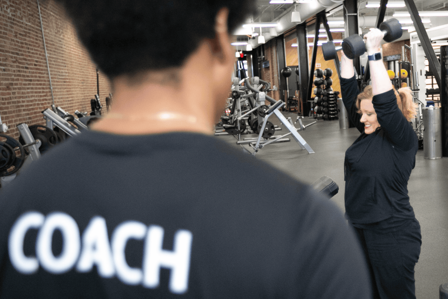 Coach observes as a woman lifts dumbbells overhead, smiling confidently during a strength training session in a gym.
