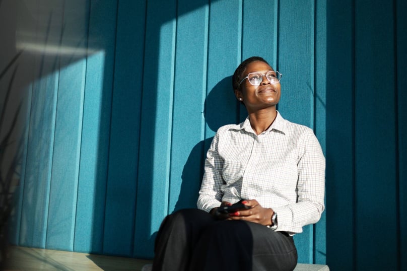 Woman in glasses sitting against a blue wall, smiling as she looks up, enjoying a moment of reflection in natural sunlight.