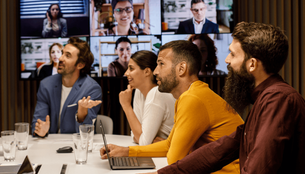 Hybrid team in a conference room engaged in a video call with global colleagues — reflecting the complexity of supporting a dispersed workforce.