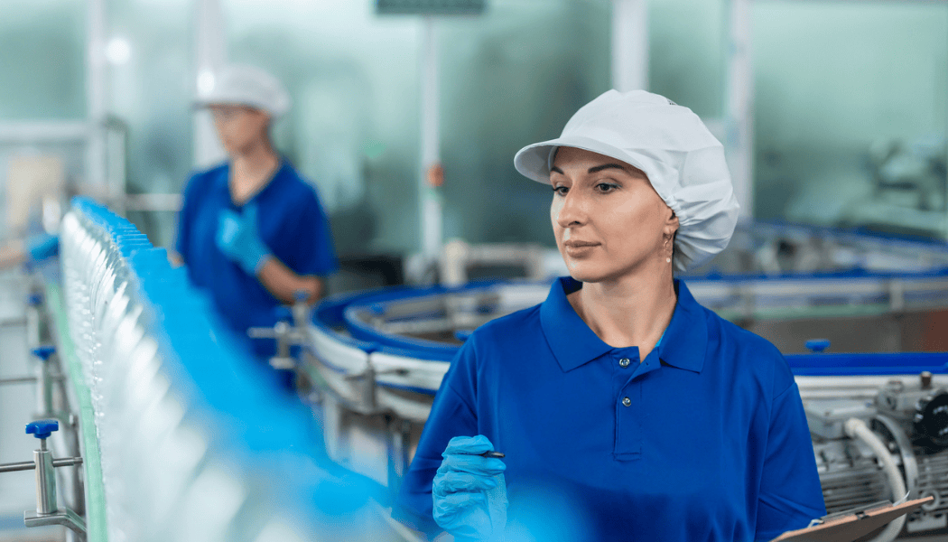 Woman in blue uniform and hairnet inspects bottled water on a production line in a factory.