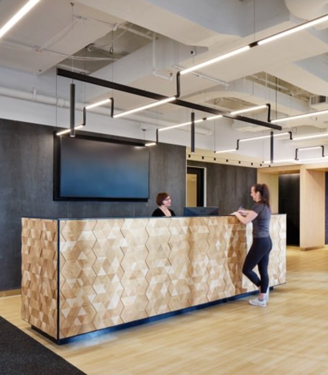 Reception area in a modern wellness facility, where a woman chats with staff while another uses a beverage station.