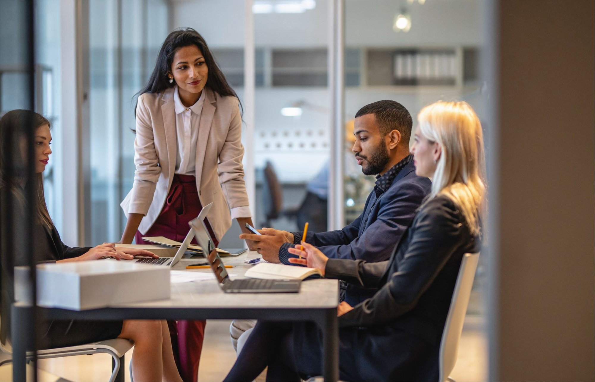 Four professionals in a modern office have a focused team discussion around a table with laptops and notebooks.