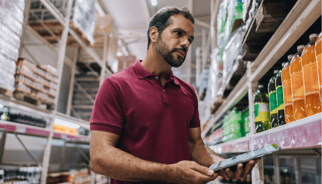 A frontline retail worker in a grocery store aisle uses a tablet to assess products, reflecting the challenges of supporting well-being in dispersed teams.