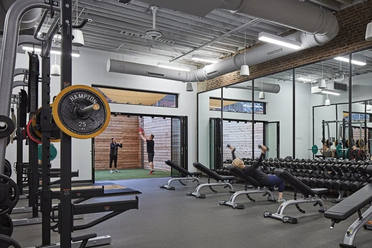 Strength training area with benches, dumbbells, squat racks, and people exercising indoors and outdoors.