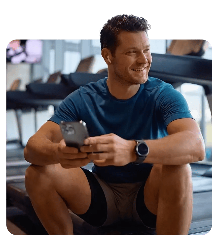 A man in workout gear sits in a fitness center holding a phone while smiling.