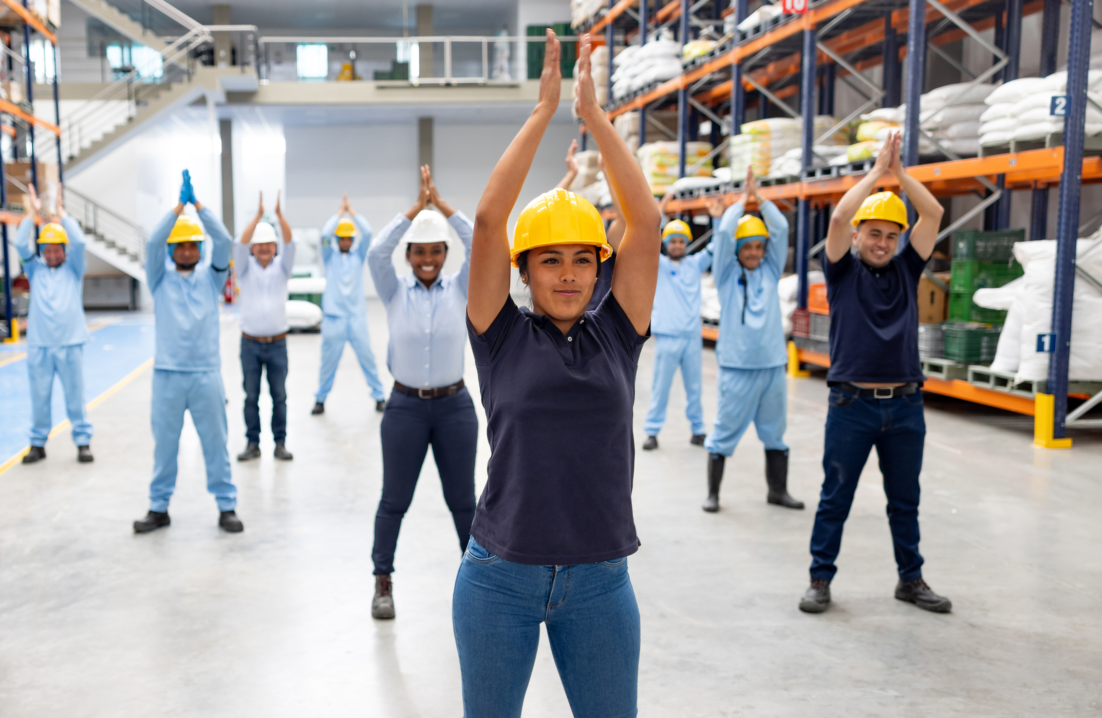 Warehouse workers in safety helmets and uniforms performing group stretching exercises together