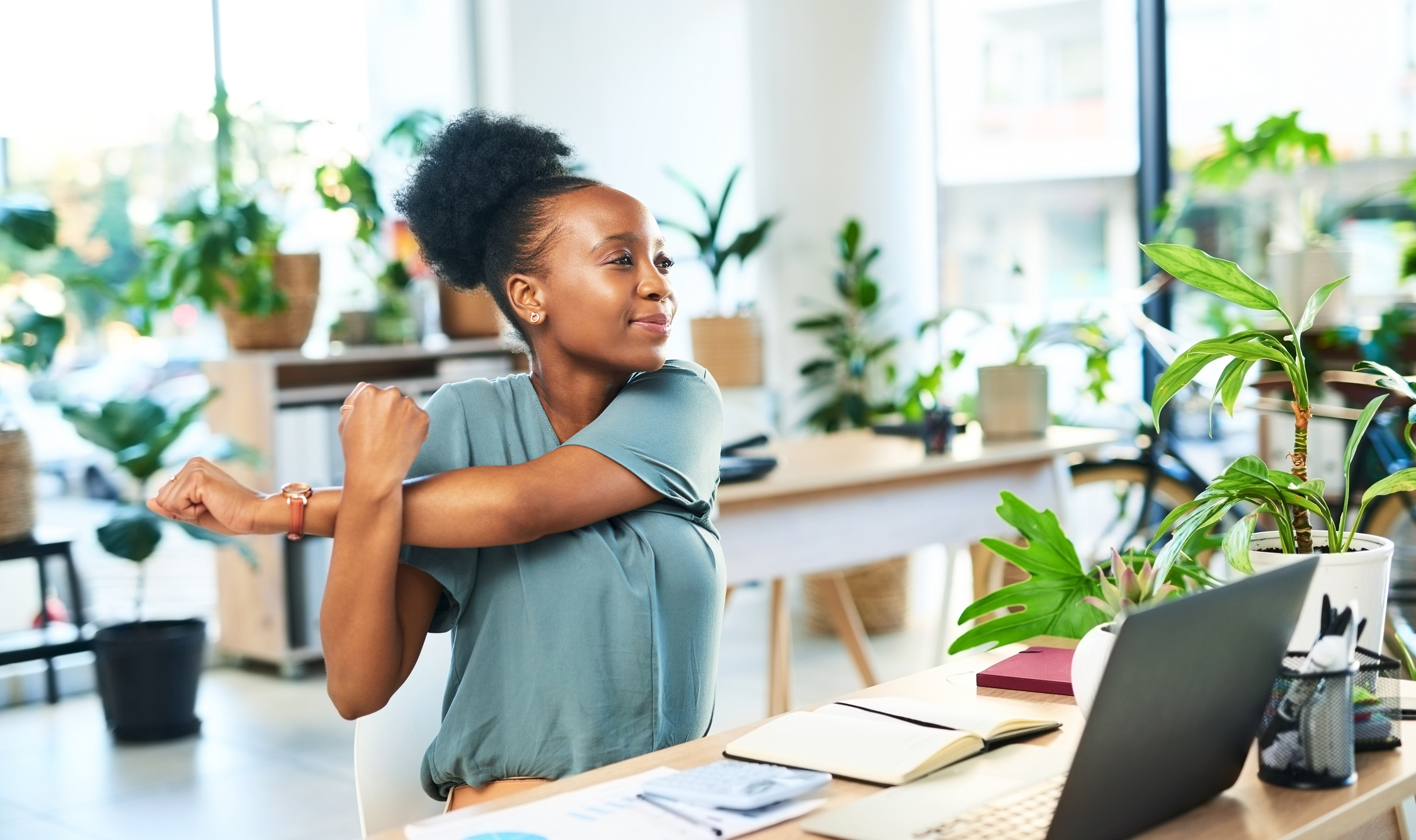 A woman in a modern office stretching her arm while sitting at a desk with a laptop, notebooks, and potted plants around her.