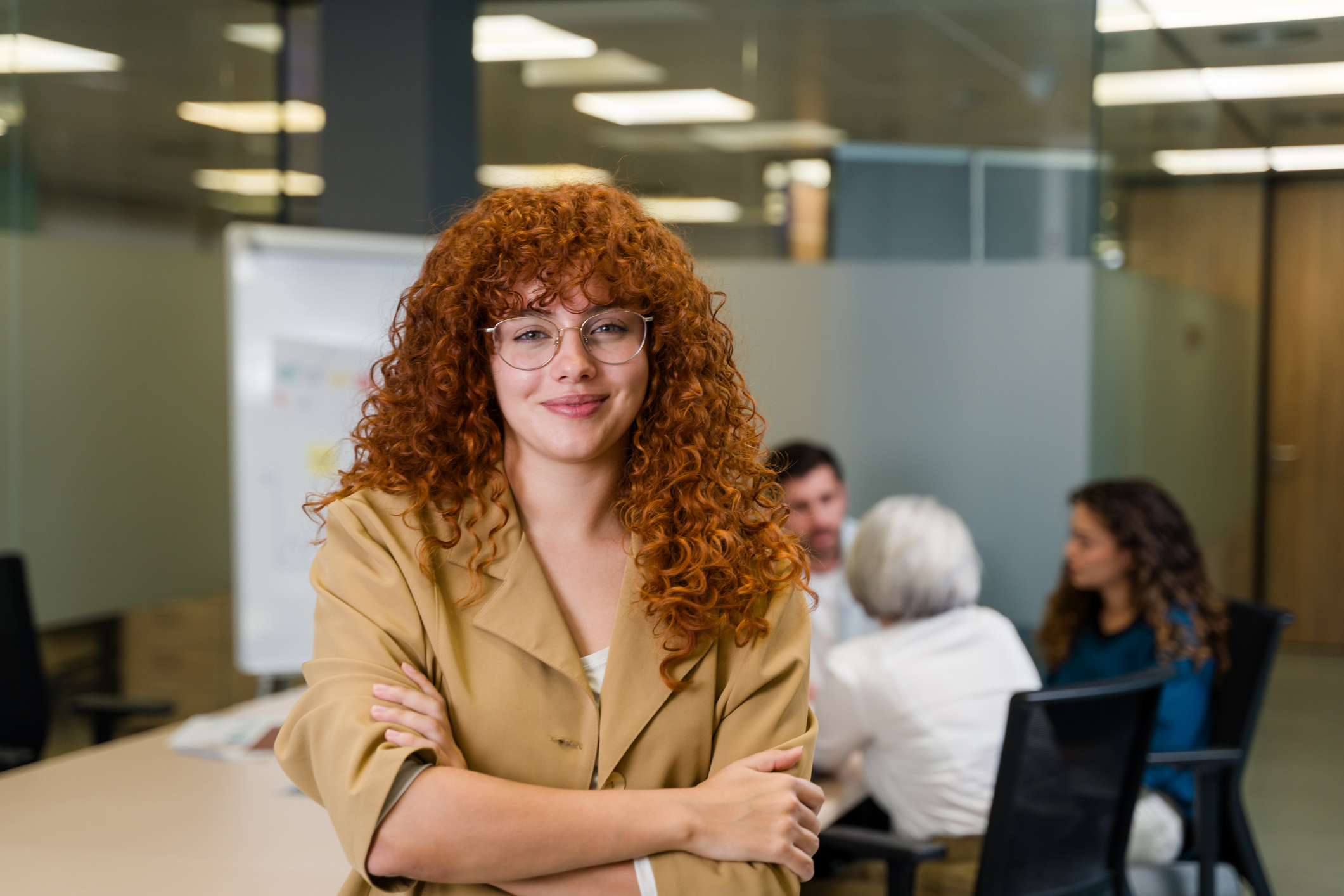 Confident young professional standing in a modern office, smiling while colleagues collaborate in the background.