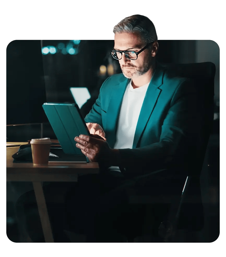 A man wearing glasses and a suit looks at his tablet in his office at night.