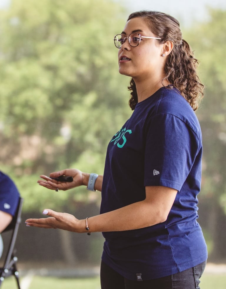 Exos coach presents to a group, holding a clicker and gesturing while speaking in a natural setting.