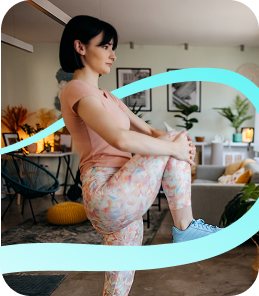 Focused woman in colorful leggings performs a standing knee hug stretch at home, surrounded by plants and cozy decor.